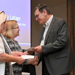 Gentleman in suit presenting an envelope to a lady and shaking her hand. Another lady stands next to her, looking down at a piece of paper.