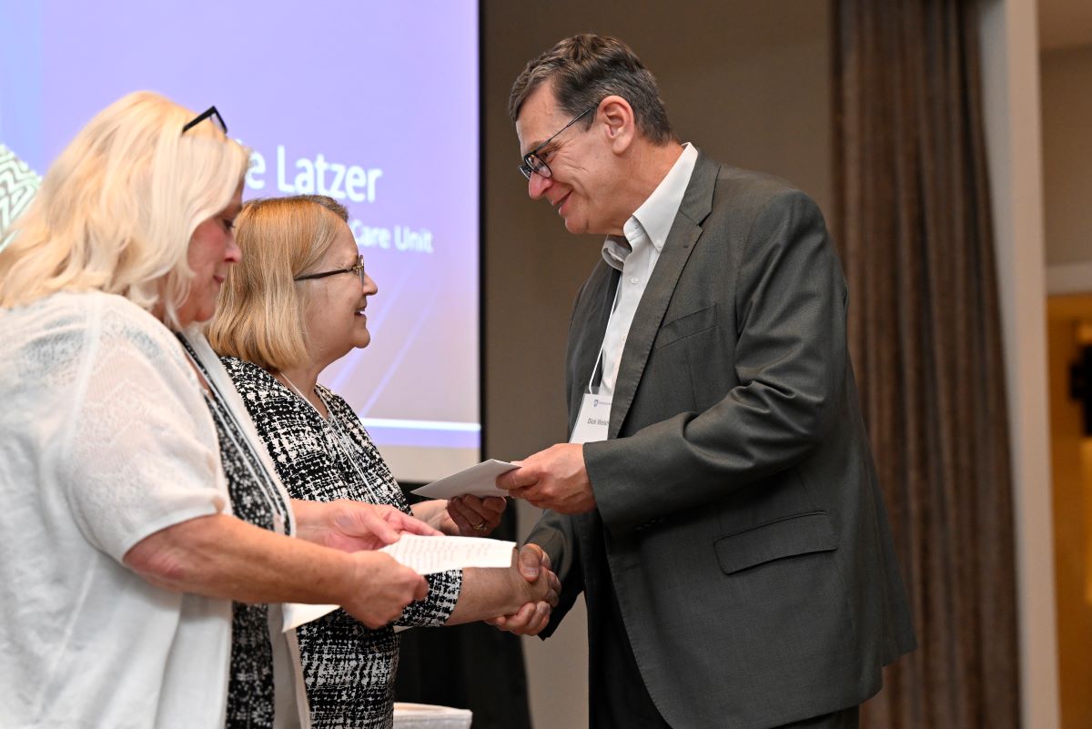 Gentleman in suit presenting an envelope to a lady and shaking her hand. Another lady stands next to her, looking down at a piece of paper.