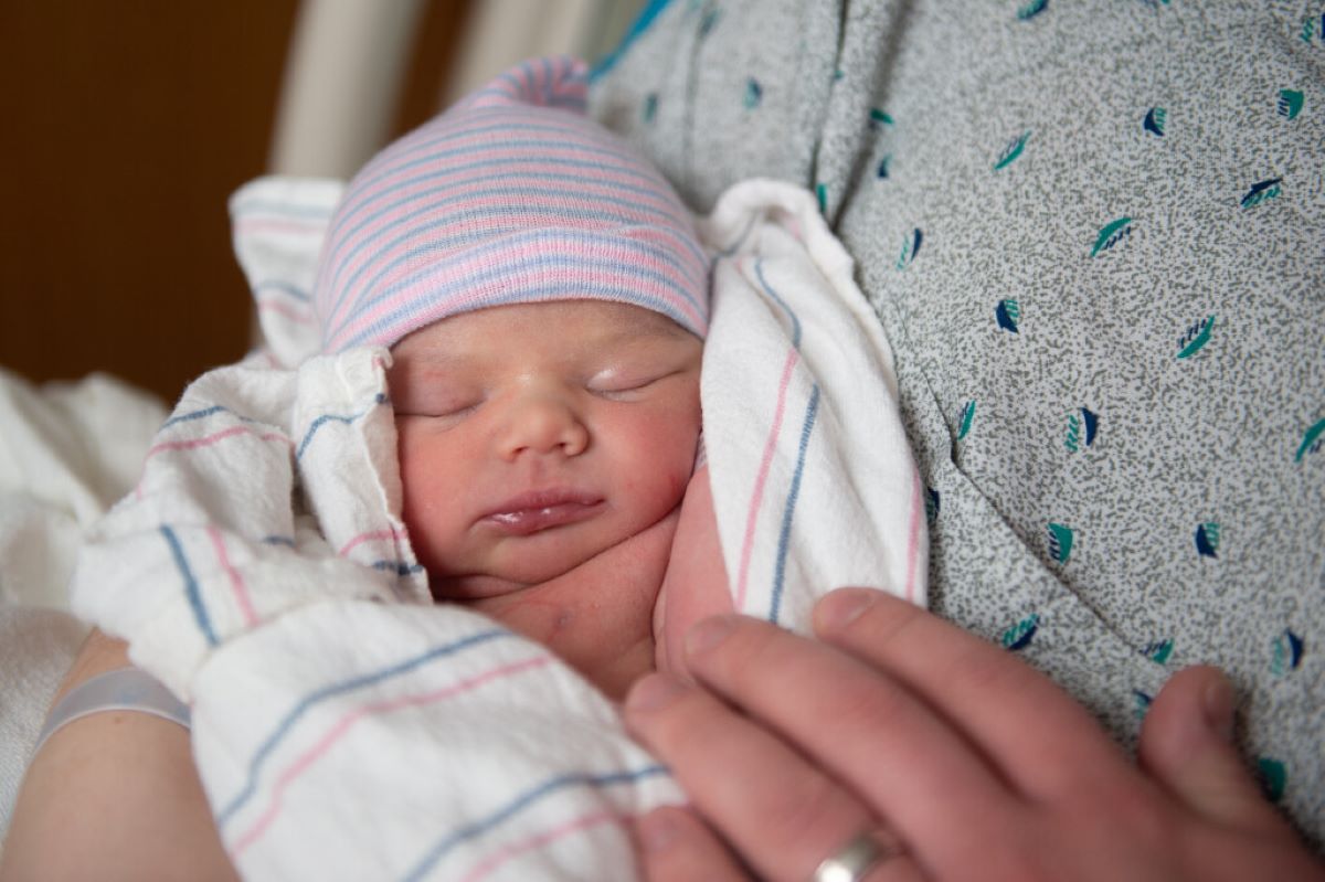 A sleeping newborn baby wearing a knit hat and swaddled in a blanket is held closely by her mother.