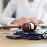 Stethoscope and gavel laying on wooden desk. Person in physician’s coat sitting at desk in blurred background.