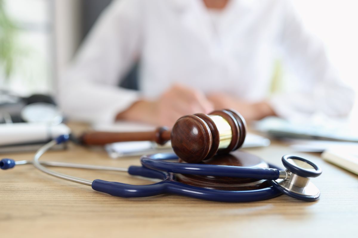 Stethoscope and gavel laying on wooden desk. Person in physician’s coat sitting at desk in blurred background.