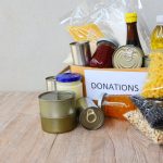 Food in cans, jars and baggies on a wooden table and in a small cardboard box with a label that says, “donations.”