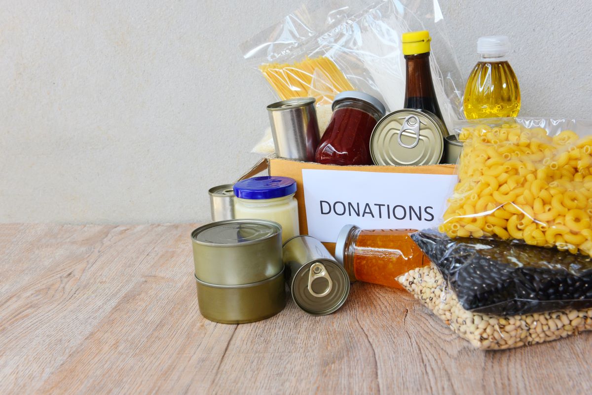 Food in cans, jars and baggies on a wooden table and in a small cardboard box with a label that says, “donations.”