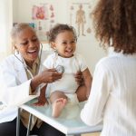 A doctor holds a stethoscope to a laughing baby while another woman looks on in a doctor's office.