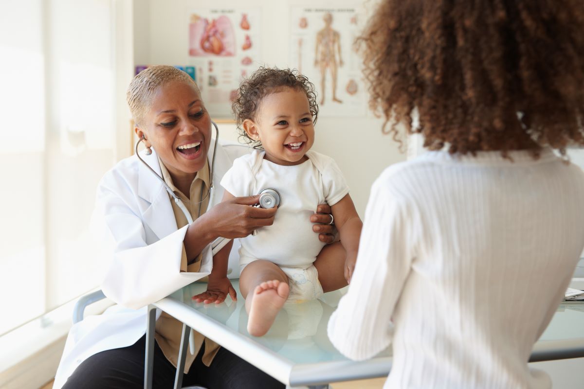 A doctor holds a stethoscope to a laughing baby while another woman looks on in a doctor's office.