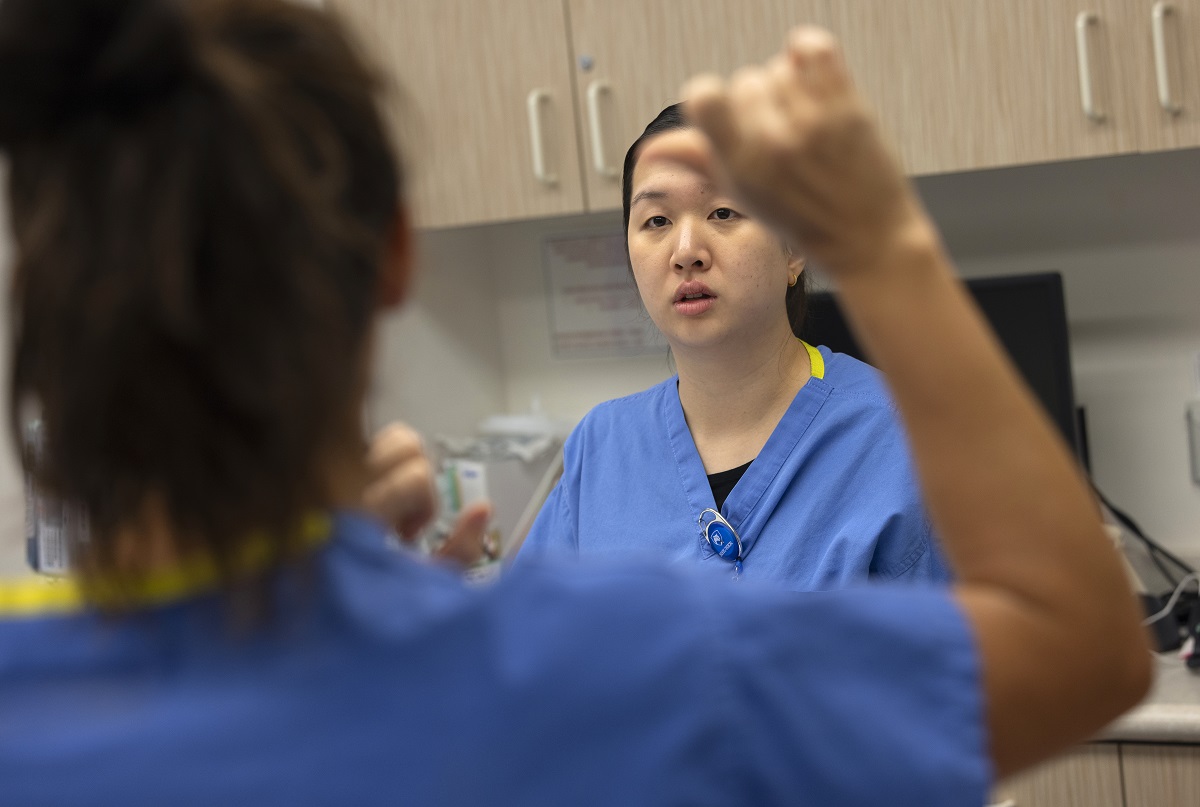 A woman in the foreground gestures at another woman wearing medical scrubs in the background.