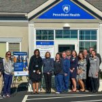 A group of men and women line up in front of a medical office building. A poster wishing staff members a happy anniversary sits on an easel between the third and fourth person from the left. The poster includes a photo of the Nittany Lion from the building’s opening celebration.