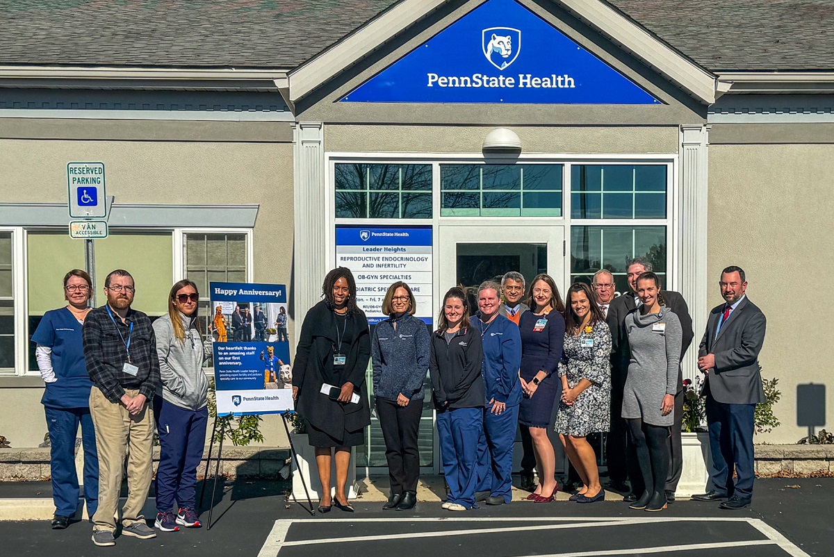 A group of men and women line up in front of a medical office building. A poster wishing staff members a happy anniversary sits on an easel between the third and fourth person from the left. The poster includes a photo of the Nittany Lion from the building’s opening celebration.