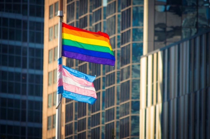 The rainbow Pride flag waves in the wind above the Transgender Pride flag, which has blue, pink and white stripes.