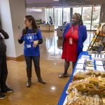 Three ladies standing in a hallway of a business building, the lady in the middle speaking to the lady on the left. There is a table to the right against a wall, displaying flyers, a poster and packaged items.