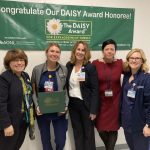 Five women stand in a row in front of a sign congratulating "Our DAISY Award winner."
