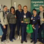 Six ladies wearing scrubs stand side-by-side in a medical facility, one holding a certificate and a gift bag. A sign is in the background that says, “Congratulate Our DAISY Award Honoree.”