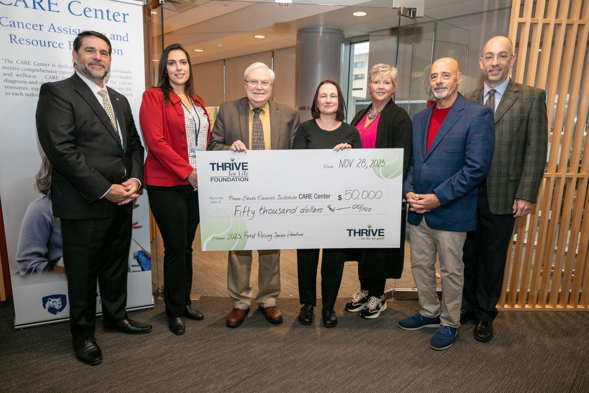 Seven people in business attire standing in a row, smiling. Two people in the middle holding a giant check for $50,000 made out to “Penn State Cancer Institute CARE Center” with the THRIVE for Life Foundation logo on it.