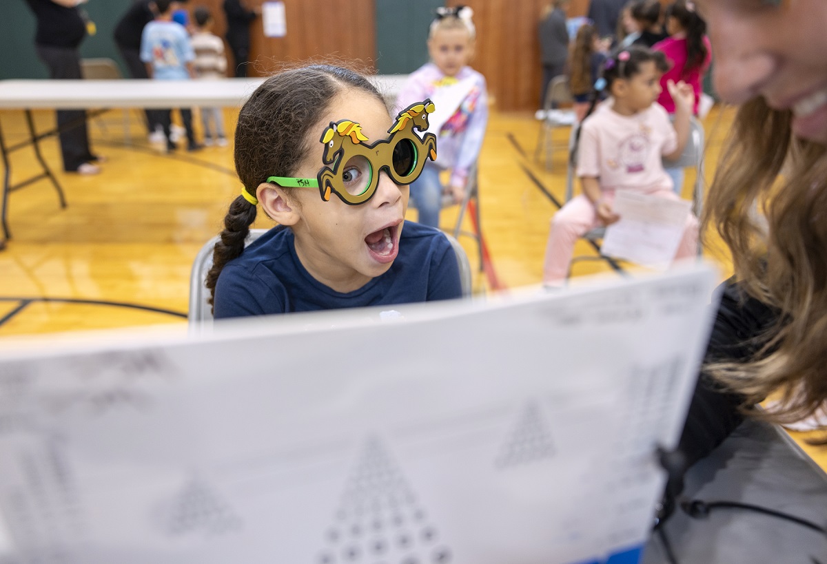 Yasmely Rodriguez, a first-grader at Henry Houck School in Lebanon, opens her mouth wide and smiles as she sits at a monitor. She is wearing glasses in the shape of horses and a T-shirt and has her hair in a braid. Behind her, several children sit at tables in a gym.