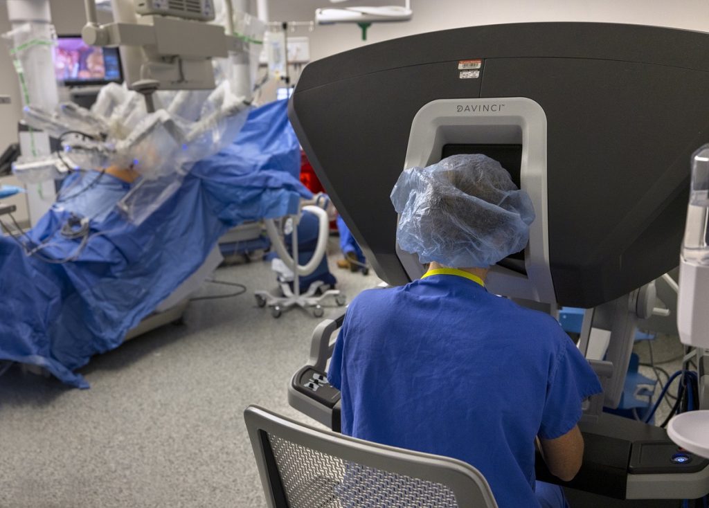 Dr. Allison Barrett is sitting with her back to the camera as she operates a da Vinci Surgical System while conducting weight loss surgery. She is wearing scrubs and a surgical cap.