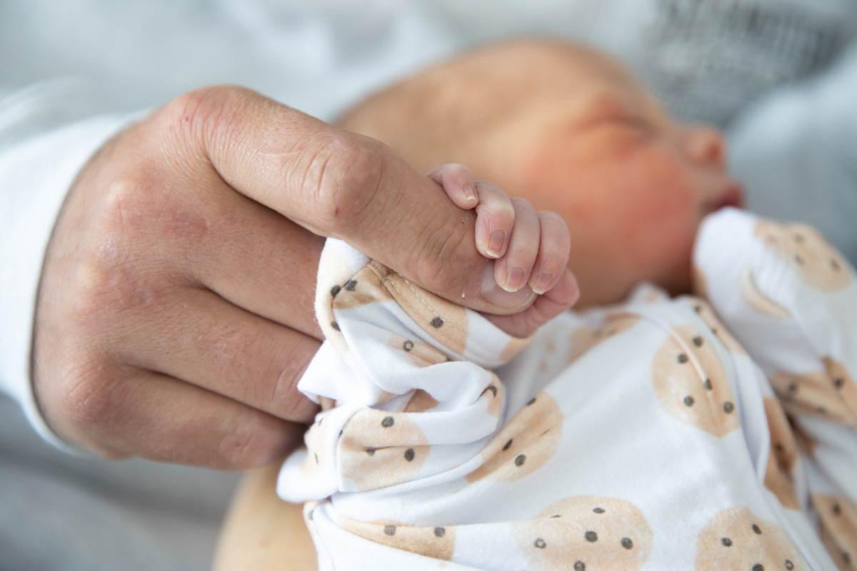 Closeup of a newborn in a onesie being held, and the baby’s right hand is wrapped around an adult finger.