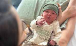 A woman holds a newborn baby in her arms. The baby is wearing a form-fitting hat and a shirt with the words "Rowan William."