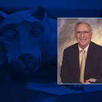 Portrait of gentleman wearing glasses, smiling, wearing a business suit; on a background of a photo of a Nittany Lion statue.