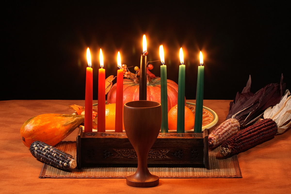 On a wooden table, a kinara holds seven lit candles, a wooden goblet in front, dried corn, pumpkin and oranges behind on the table.