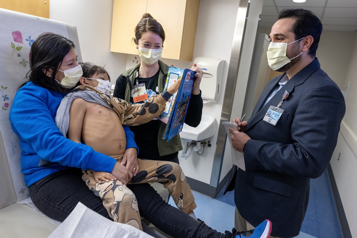 Four people in a hospital room talk behind surgical masks. One is a child, who sits on a woman's lap. The child's shirt has been pull aside. He gestures at a man who stands taking notes on a pad.