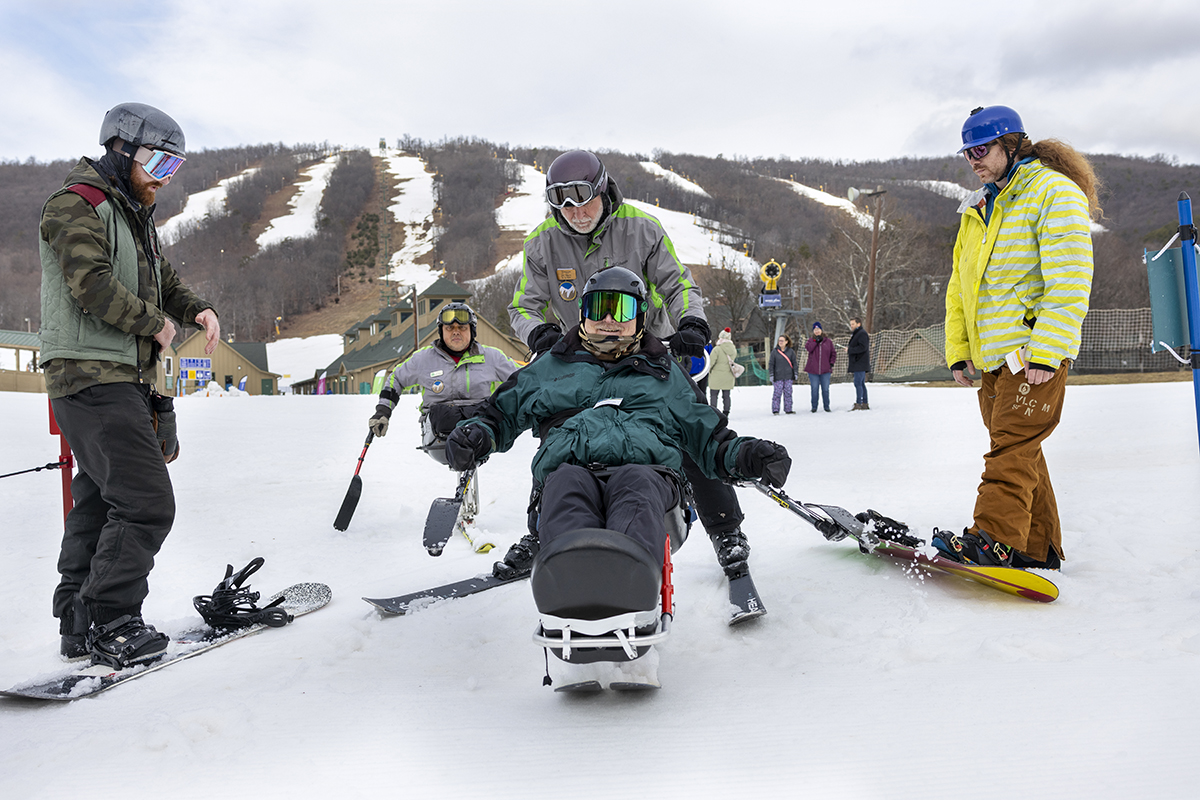 A group of people in parkas riding skis gather at the base of a mountain striped with ski runs. One is seated on a sit-ski, a device that balances his body on a single runner, and his legs are propped in front of him.