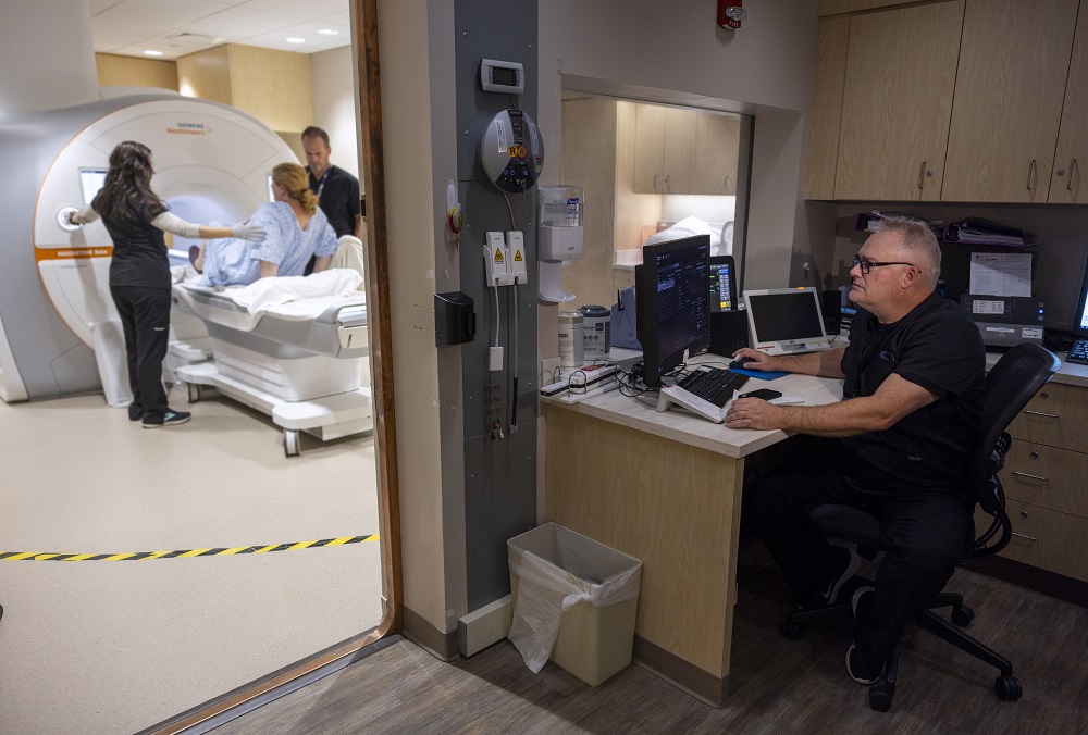 Fred Gajewski, right, sits at a desk and operates a computer to perform an MRI. He is wearing a polo shirt and glasses. In the room to his left, two technologists assist a patient in lying down before entering the MRI machine for a scan.