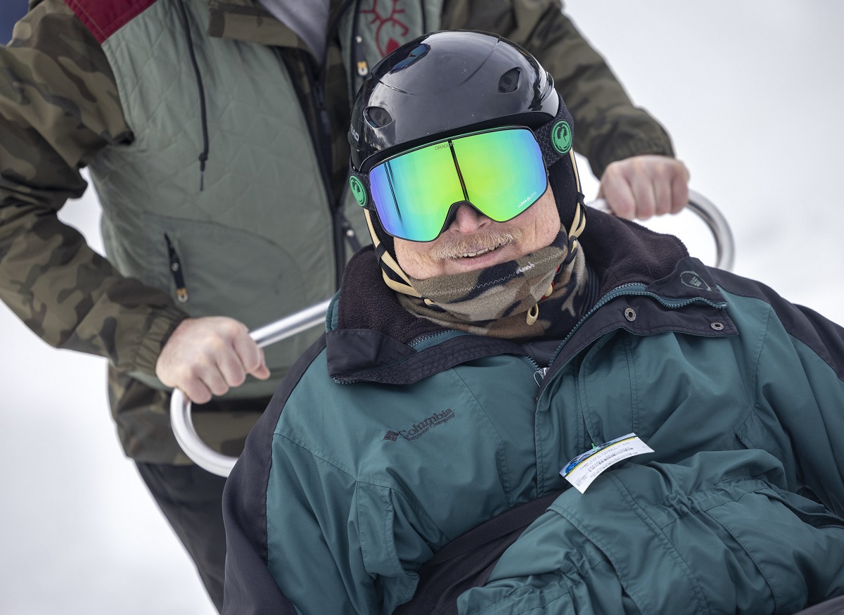 Close up of a man in ski goggles and a helmet. Beneath his mustache, he's smiling. Behind him, a pair of hands grip the handle of his sit ski.