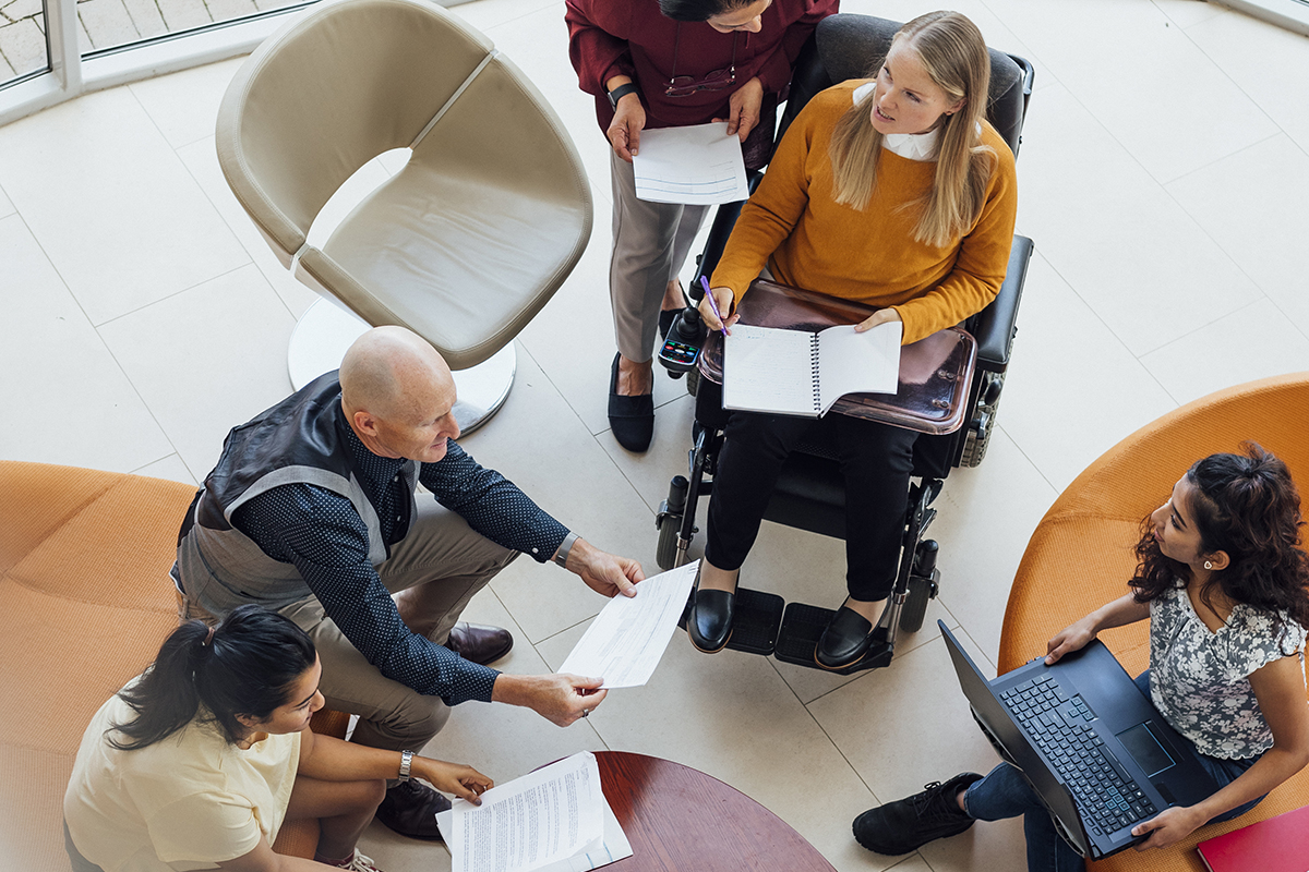 Four people sitting in a circle, one standing, holding laptops and papers. One person sits in a wheelchair.