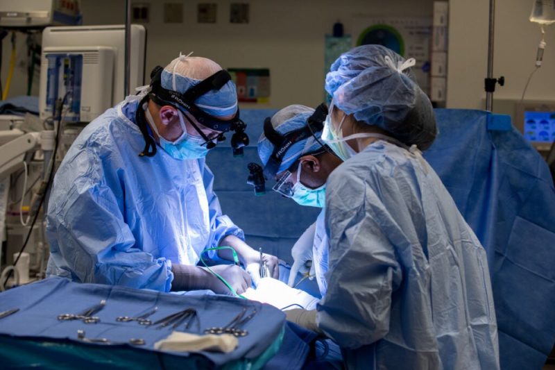 Three medical professionals in surgical gear operate on a patient, not shown. A tray of surgical instruments is within easy reach.
