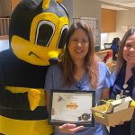 A female hospital employee wearing scrubs and holding a certificate, stands between a person dressed in a bumble bee costume and a female nurse wearing hospital scrubs.