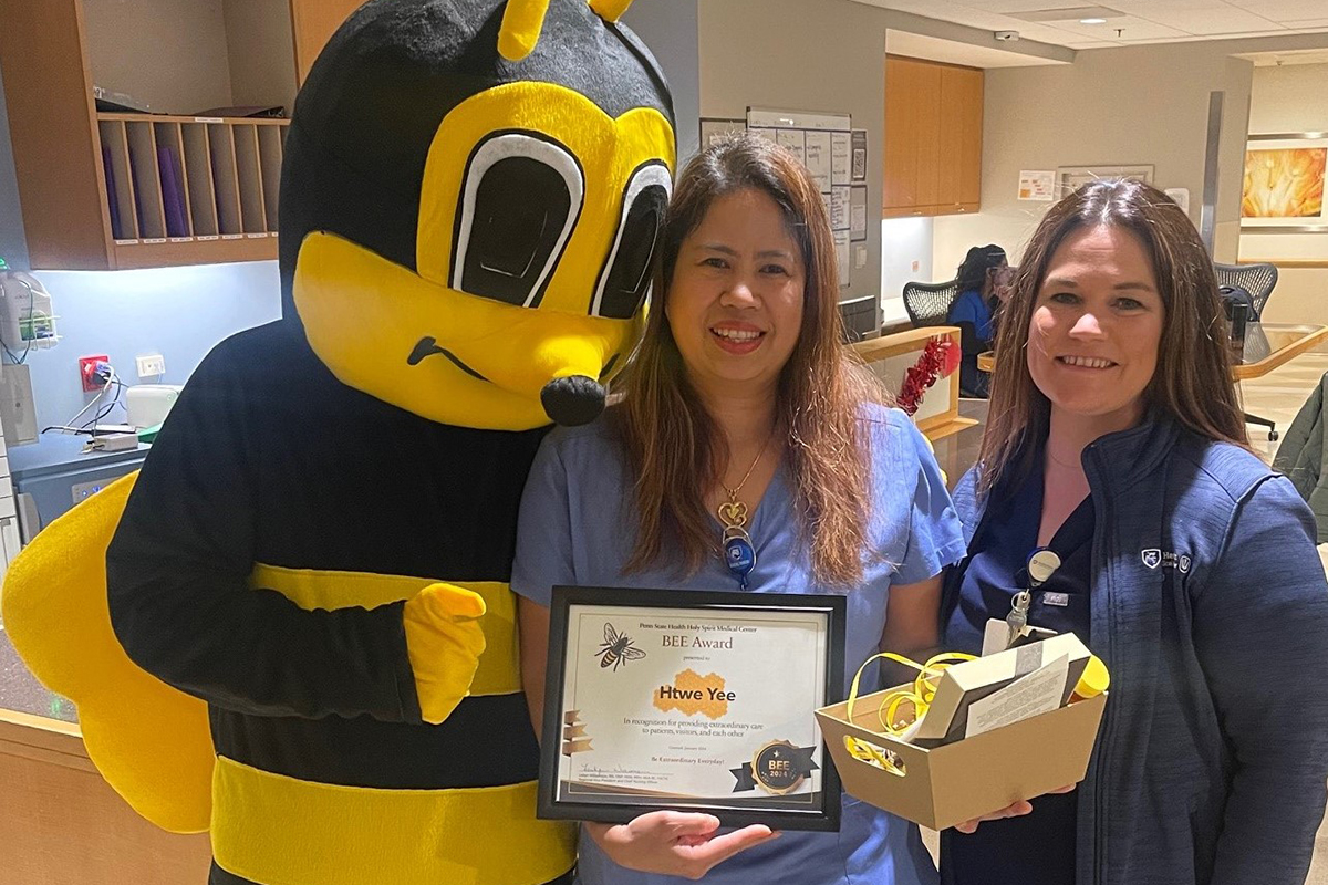 A female hospital employee wearing scrubs and holding a certificate, stands between a person dressed in a bumble bee costume and a female nurse wearing hospital scrubs.