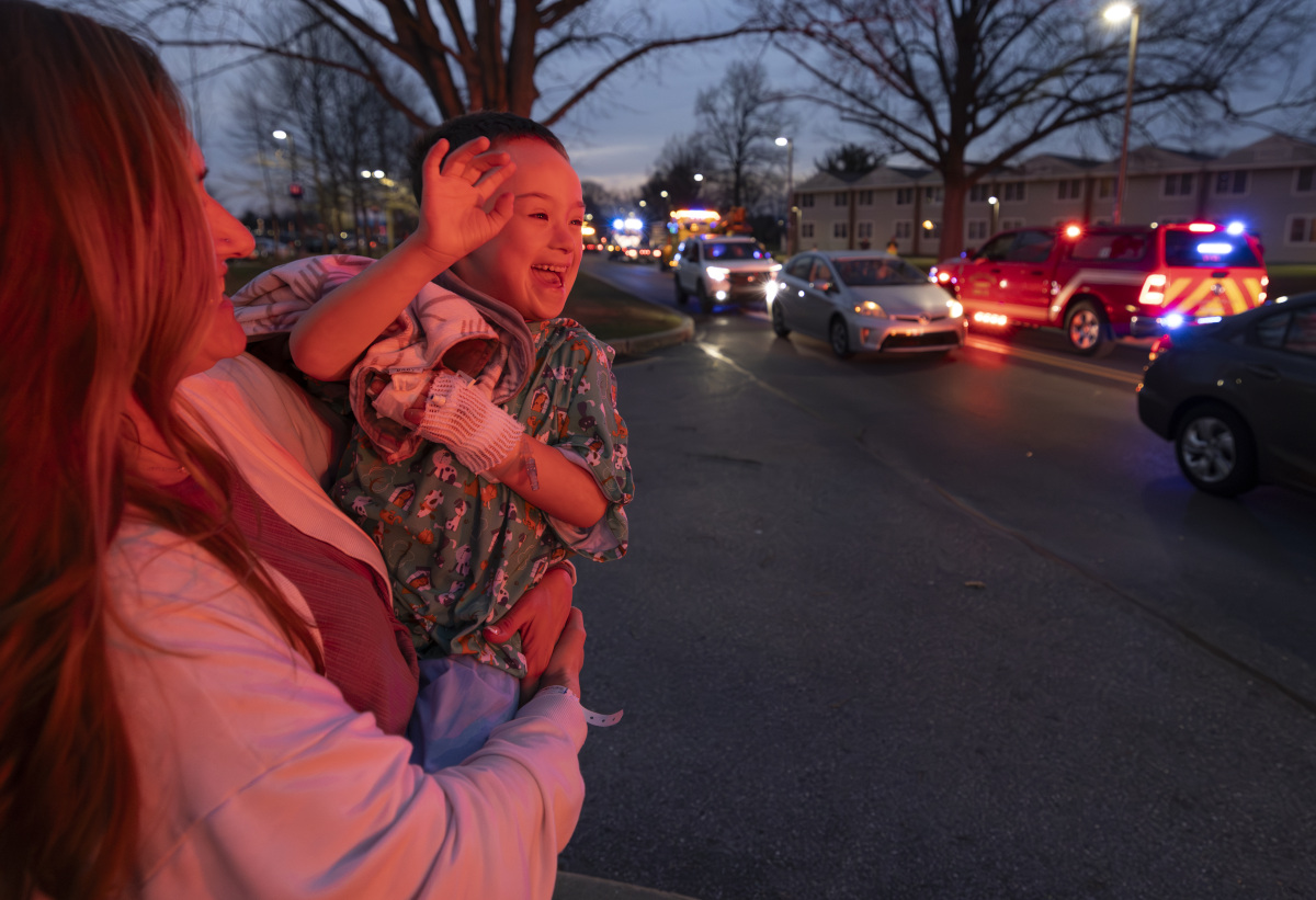 A woman holds a young boy in her arms. He is smiling and waving to several emergency vehicles as they drive by with their lights flashing.
