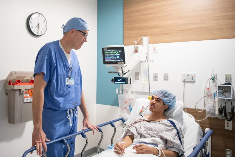 A doctor dressed in surgical scrubs stands next to the bedside of a patient. They are talking.
