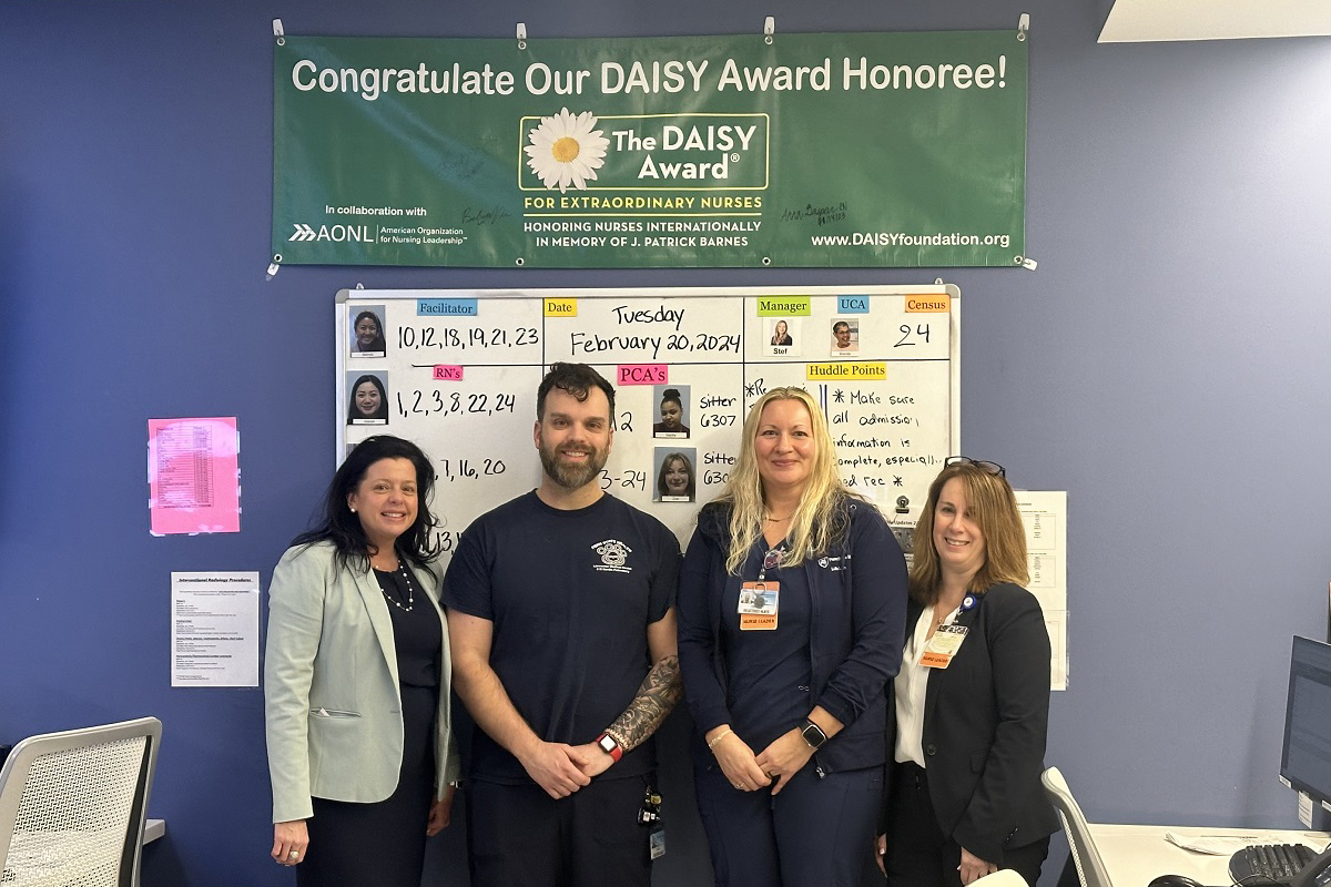 Four people stand side-by-side, smiling against a wall with a marker board, and a sign that says, “Congratulate Our DAISY Award Honoree! The DAISY Award for extraordinary nurses. Honoring nurses internationally in memory of J. Patrick Barns.”