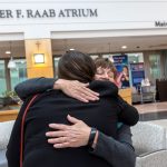 Two women hug in the lobby of Holy Spirit Medical Center. One woman is seen from behind wearing a black jacket. A sign with the words, “Walter F. Raab Atrium” is above them.