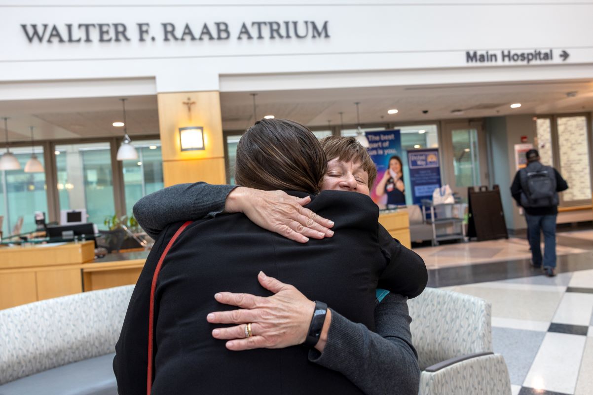 Two women hug in the lobby of Holy Spirit Medical Center. One woman is seen from behind wearing a black jacket. A sign with the words, “Walter F. Raab Atrium” is above them.