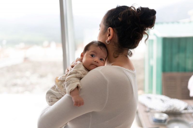 A woman holding a baby – the woman faces away looing out a window with the baby facing over her shoulder.