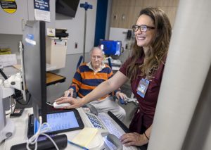 A female registration associate, wearing glasses and maroon scrubs, smiles while working on a computer tablet in a hospital emergency room, with a male patient seated in the background, wearing a striped shirt.