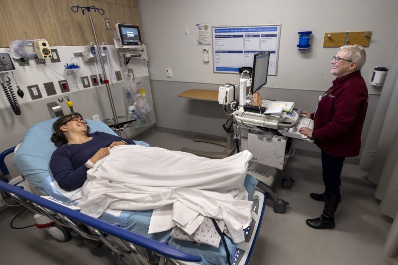 In a bright hospital emergency room a female patient, wearing glasses and a blue shirt, is lying in a bed, covered by a white blanket. A smiling patient registration associate in a burgundy sweater stands at a mobile computer station to the right, amidst various medical devices.
