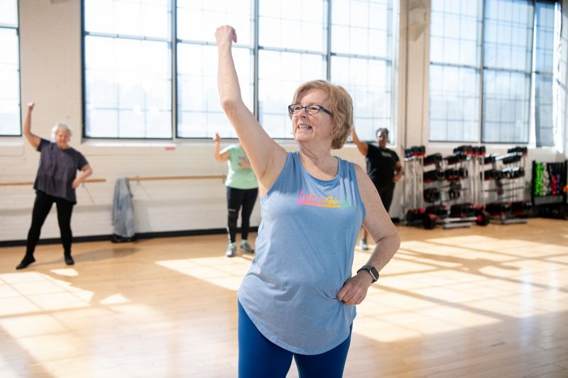 Patricia Creque puts her right arm in the air as she leads a dance class. She is wearing glasses, a T-shirt and tights. Behind her are three women, racks with hand weights and large windows.