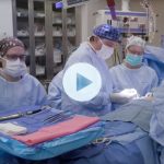 Four people in scrubs and surgical masks around a patient in an operating room while the surgeon in the center operates