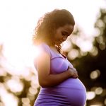 Pregnant woman standing outside, holding one hand on her tummy, looking down at it with a contented expression.