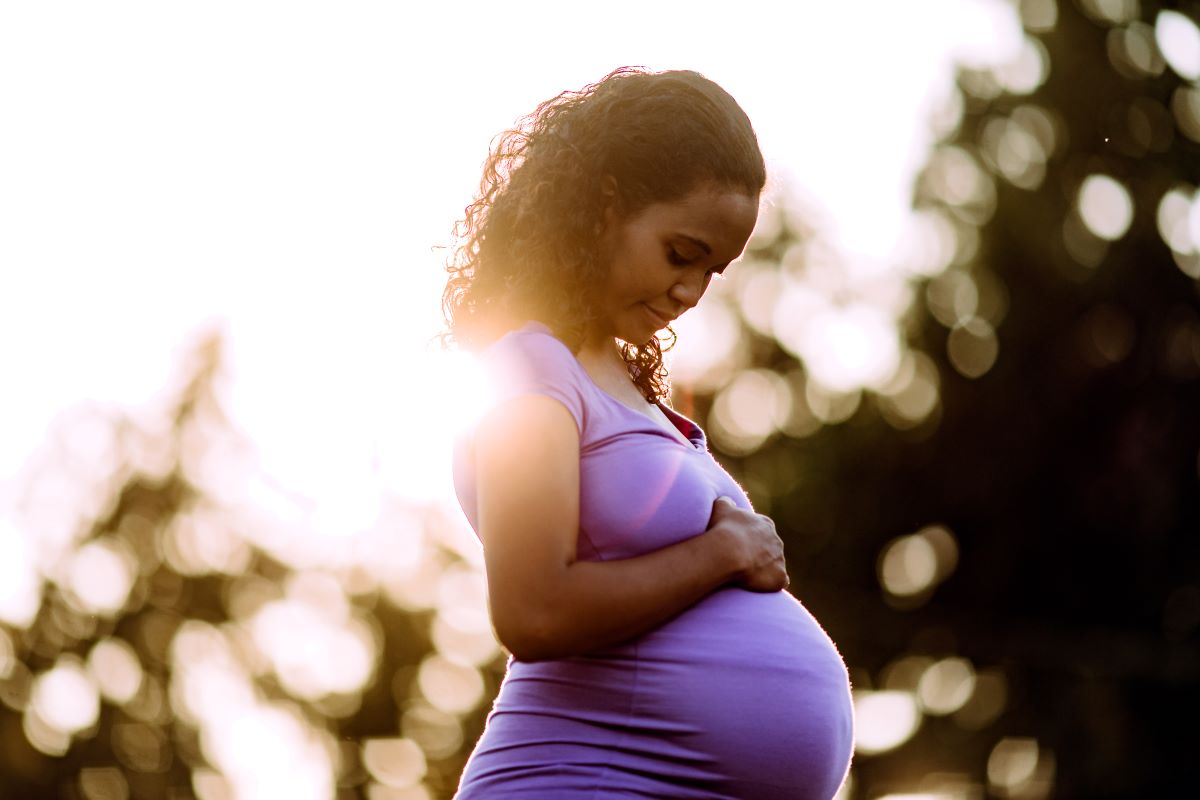 Pregnant woman standing outside, holding one hand on her tummy, looking down at it with a contented expression.