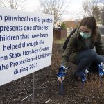 Dr. Kathryn Crowell crouches down as she puts a pinwheel in the ground in front of Penn State Health Milton S. Hershey Medical Center in 2023. A sign to her left says: “Each pinwheel in this garden represents one of the 579 children that have been helped through the Penn State Hershey Center for the Protection of Children during 2022.”
