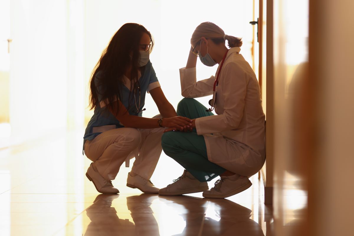 Two female hospital staff members squat on the floor in the hallway of a medical facility. One leans against the wall with her hand on her head. The other holds her hand.