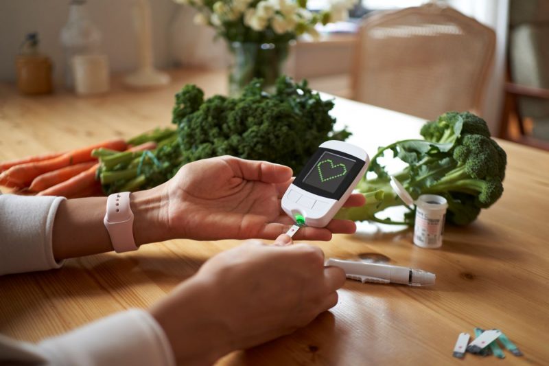 A woman’s hands, resting on a table, are holding a diabetes monitor into which she has inserted a test strip. Next to her hands are additional testing strips and a lancet holder. To the left of her hands are carrots and kale and to the right of them broccoli. In the background, out of focus, are a vase of flowers and a small dining room-style table. The monitor she is holding displays a heart.