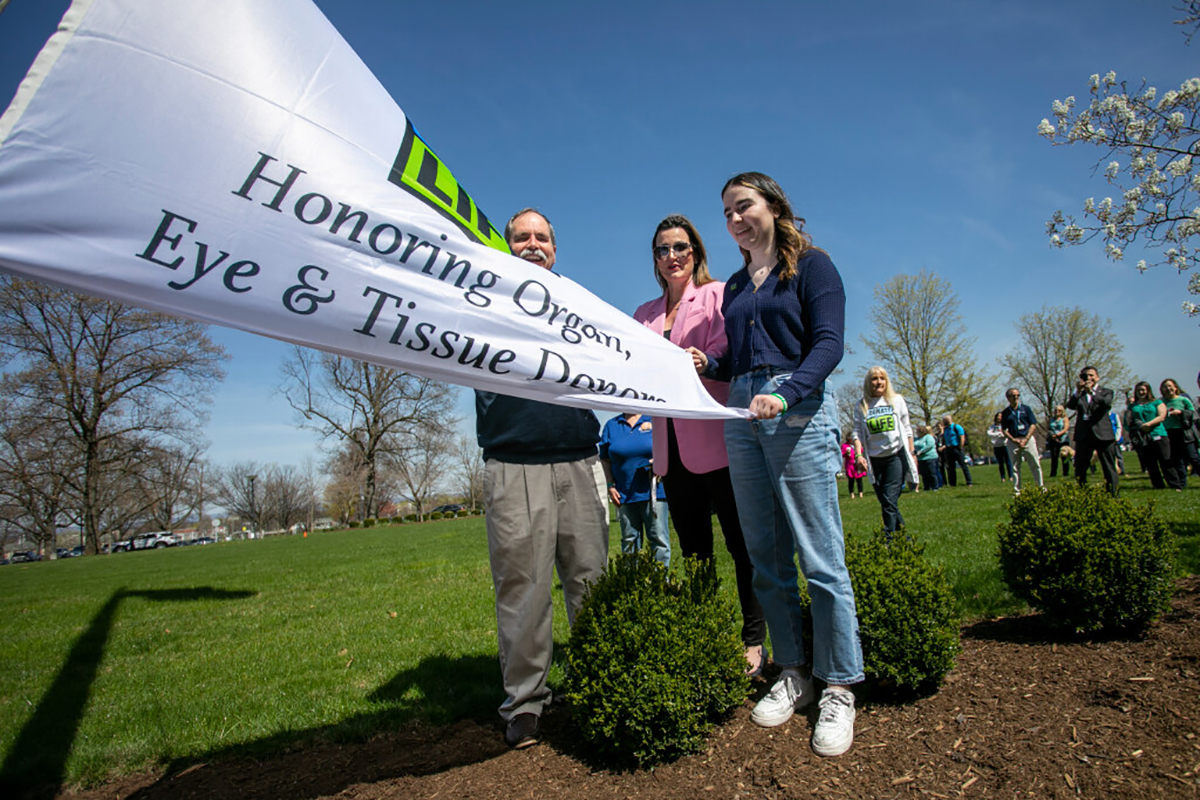 A flag marked with the words Honoring Organ, Eye and Tissue Donors is held in the center by Greg Allen, dressed in pants and a sweater. Along with his two brothers, who are not pictured, he received a heart transplant. To the right of him are Kellie Persofsky, who is wearing pants and a blazer, and her daughter, Sophia, dressed in jeans and a sweater. Mrs. Persofsky donated her kidney to her daughter. In the background are a number of other people who are taking part in the 2023 Donate Life flag-raising ceremony at Penn State Health Milton S. Hershey Medical Center. The event honors organ donors and recipients.