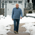 Gentleman walks on a neighborhood sidewalk wearing jeans and a sweater. A house is in the background. Snow covers the yard.