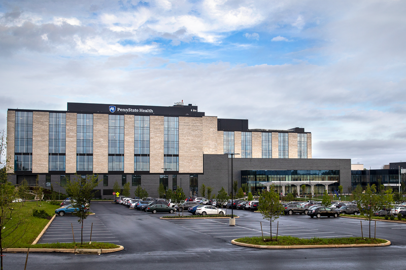 The front of a hospital building with a parking lot in the foreground and a mostly cloudy sky above. The building has a sign on the front that has the Penn State Nittany Lion shield and “Penn State Health.”