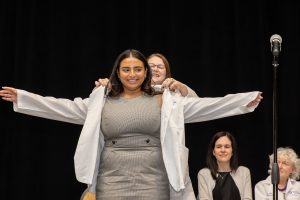 A person smiles as another person puts a short white coat on them, signifying their entry into the medical profession.